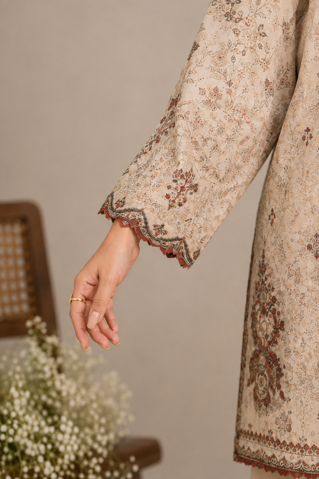 Close-up of a person wearing a beige printed kurta with brown and muted red floral motifs and scalloped embroidered cuffs, shown against a soft neutral background with a blurred chair and flowers.