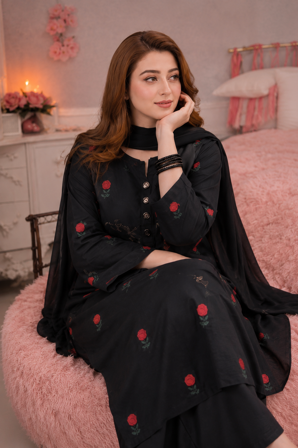 A woman wearing a black traditional outfit with red rose embroidery sits on a pink fluffy chair in a decorated bedroom setting.