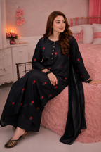 A woman models a black three-piece ethnic suit with red floral embroidery, featuring a long kurta, wide-leg pants, and a matching dupatta, posed on a pink bed in a bedroom setting.