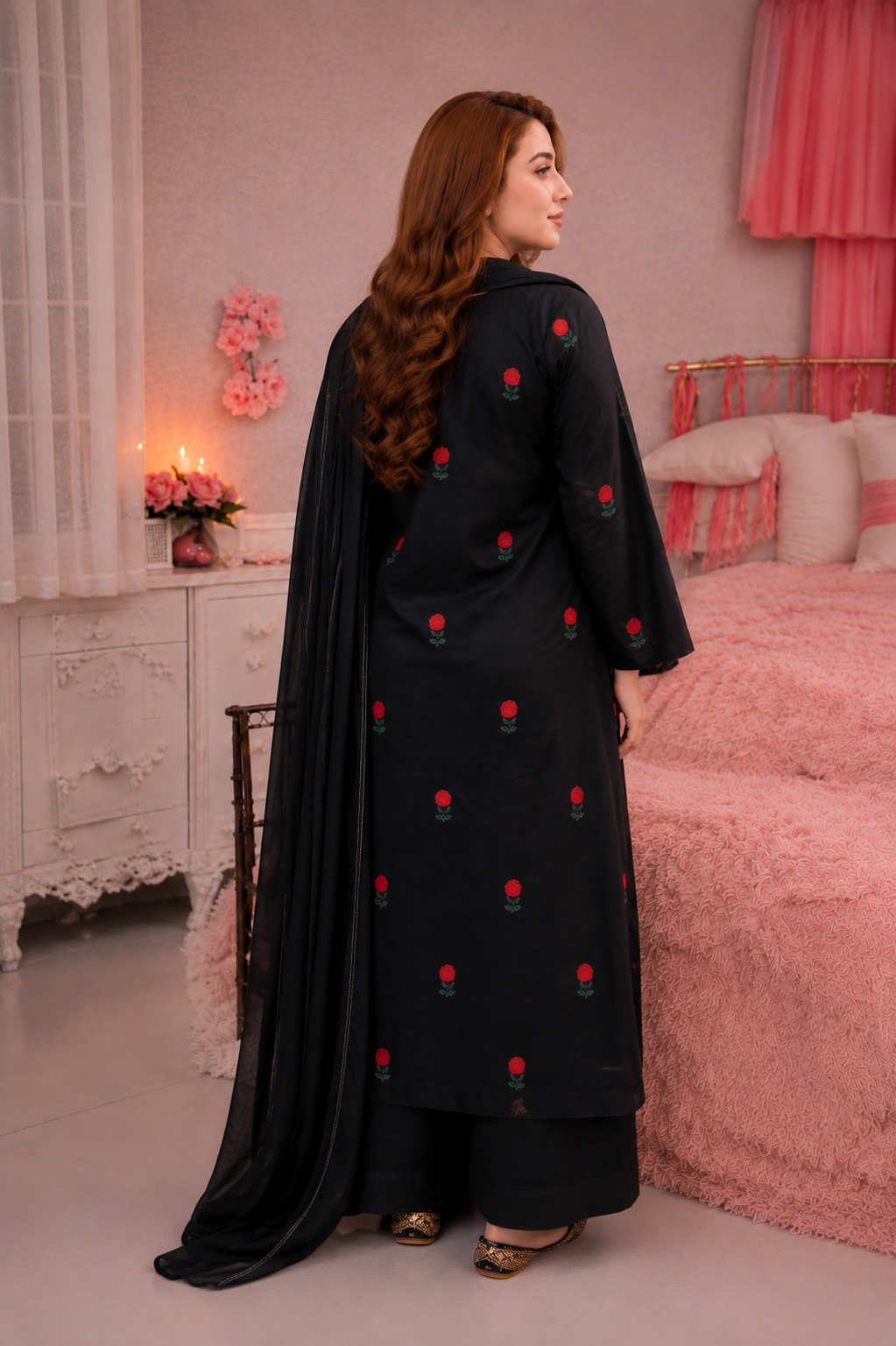 A woman wearing a black traditional outfit with red floral embroidery and a matching black dupatta, standing in a pink bedroom setting.
