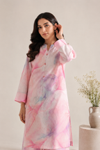 A woman modeling a light pink long kurta with a watercolor-style abstract print in pink, purple, and blue, set against a warm beige background with a small vase of dried flowers on the side.