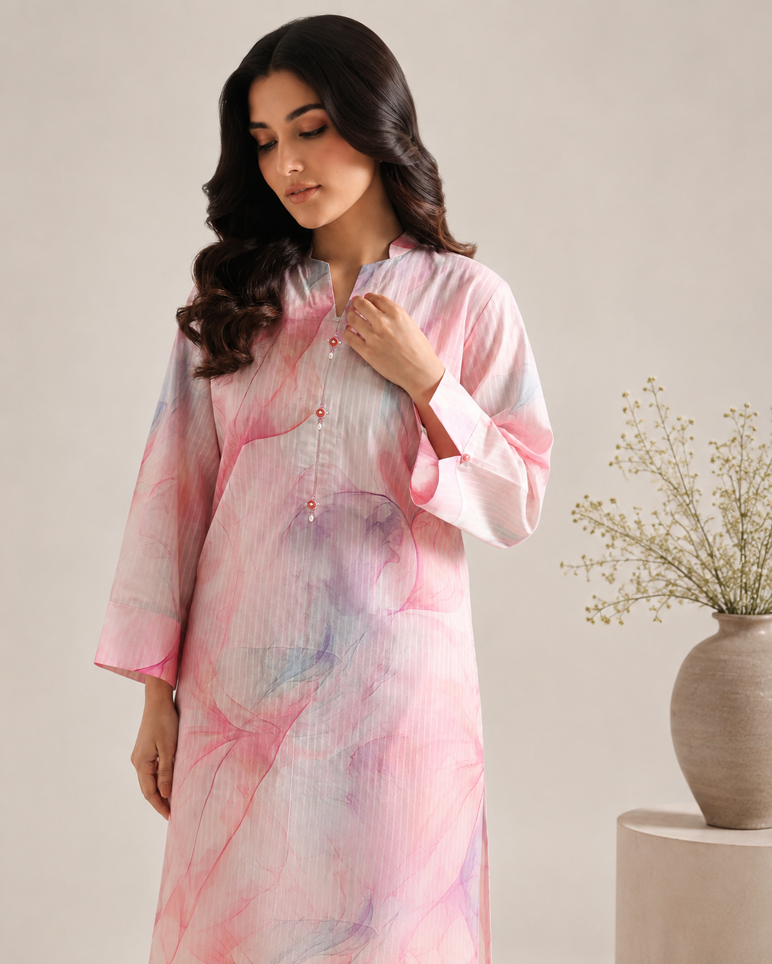 A woman wearing a long-sleeved, high-neck kurta with a pink, purple, and white abstract watercolor print, posed next to a neutral-toned ceramic vase with dried flowers.