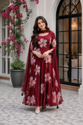 A woman models a long-sleeved maroon red anarkali gown with a pink floral print and a matching sheer cape overlay, set against an outdoor patio backdrop with a black arched window and pink flowers.