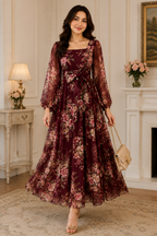 A woman models a long-sleeved, floor-length floral dress in a rich burgundy and pink floral print. The dress features a square neckline, a self-tie waist, and a flowing skirt.