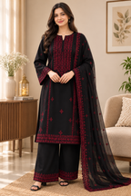 A woman models a black three-piece Indian ethnic suit with deep red embroidery, featuring a long kurta, wide-leg trousers, and a matching sheer dupatta, set against a warm, modern living room background.