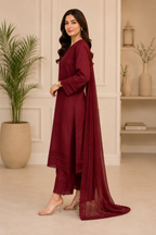 A woman models a deep maroon three-piece traditional outfit with a matching sheer dupatta in an elegant indoor setting with a potted plant and shelves.