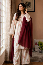 A woman models a traditional South Asian outfit featuring an off-white kurta and wide-leg trousers with pink floral embroidery, paired with a deep maroon dupatta, standing in a warmly lit room by a window.