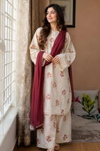 A woman stands indoors by a window wearing a cream-colored traditional outfit with red floral embroidery and a deep maroon dupatta. The setting includes a beige wall, framed wall art, sheer curtains, and a bed in the background.