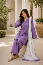 A woman models a purple three-piece salwar kameez suit with a white dupatta featuring purple borders, sitting on a stone bench in a courtyard setting.