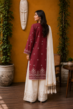 A woman wearing a traditional South Asian outfit with a maroon kurta, cream palazzo pants, and a cream dupatta, standing in front of a warm orange wall with decorative plants and a mask.