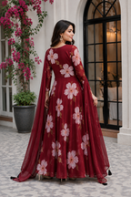 A woman wearing a long-sleeved maroon Anarkali gown with a pink floral print and a matching sheer dupatta, standing in an elegant courtyard.