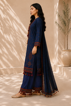 A woman modeling a navy blue traditional outfit with red and gold embroidered borders, tassel details, and a matching sheer dupatta in a sunlit indoor setting with leaf shadows.