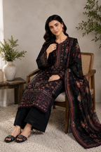 A woman seated in a wooden chair wearing a black traditional embroidered outfit with floral and geometric patterns in red, pink, and gold, paired with a matching sheer dupatta. The setting is a styled indoor room with a textured wall, a side table with a vase, and a carpet.