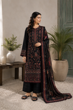 A woman modeling a black three-piece traditional outfit with intricate red and gold floral embroidery, paired with a matching dupatta, in a styled indoor setting with potted plants.