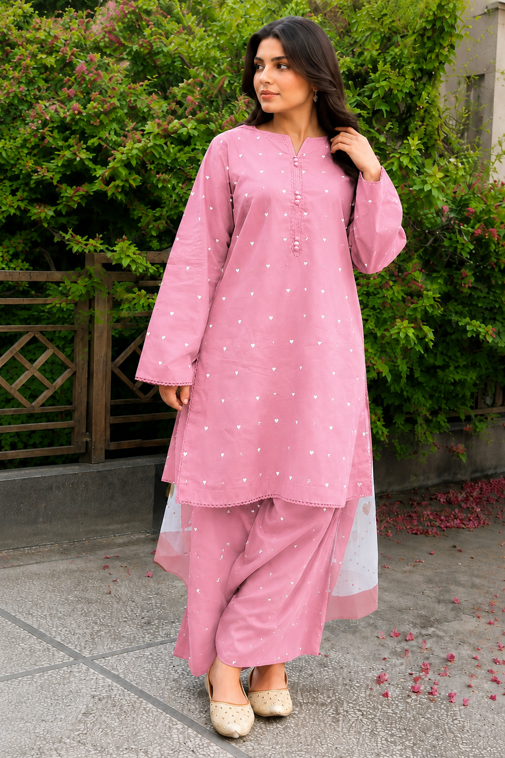 A woman wearing a pink embroidered kurta and matching palazzo pants with a white inner lining, standing outdoors in front of green bushes and a wooden fence.