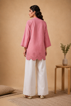 A woman stands with her back to the camera wearing a dusty pink three-quarter sleeve tunic with a scalloped hem and white wide-leg trousers, set against a warm beige studio backdrop with a small wooden side table and vase.