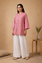 A woman modeling a pink embroidered kurta with three-quarter sleeves and white wide-leg trousers, standing in a warm beige studio setting with a small wooden side table and vase in the background.