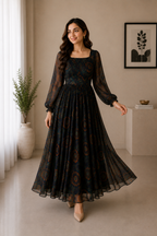 A woman modeling a long-sleeved black maxi dress with a brown and gold paisley pattern, standing in a minimalist beige-toned room.