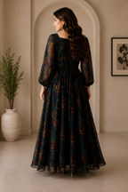 A woman stands in profile wearing a long dark teal maxi dress with a rust-orange floral pattern, photographed in a warm neutral-toned interior with an arched doorway and potted plant.