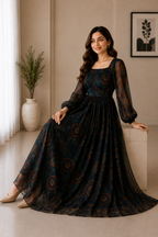 A woman models a long-sleeved black maxi dress with a brown circular mandala pattern, posed in a warm, neutral-toned indoor setting.