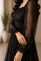 A woman wearing a dark sheer long-sleeve dress with a rust-orange floral pattern, standing indoors near a window with a potted plant in the background.