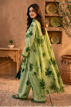 A woman modeling a light green three-piece traditional outfit with dark green floral prints, standing in a rustic indoor setting with terracotta walls and decorative plates.